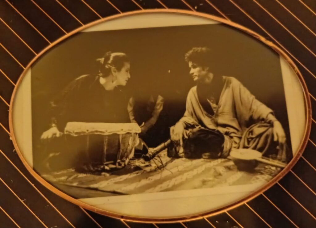 Photographie de scène en sépia dans un cadre ovale : une comédienne penchée sur un tambour face à un musicien assis en tailleur sur un tapis